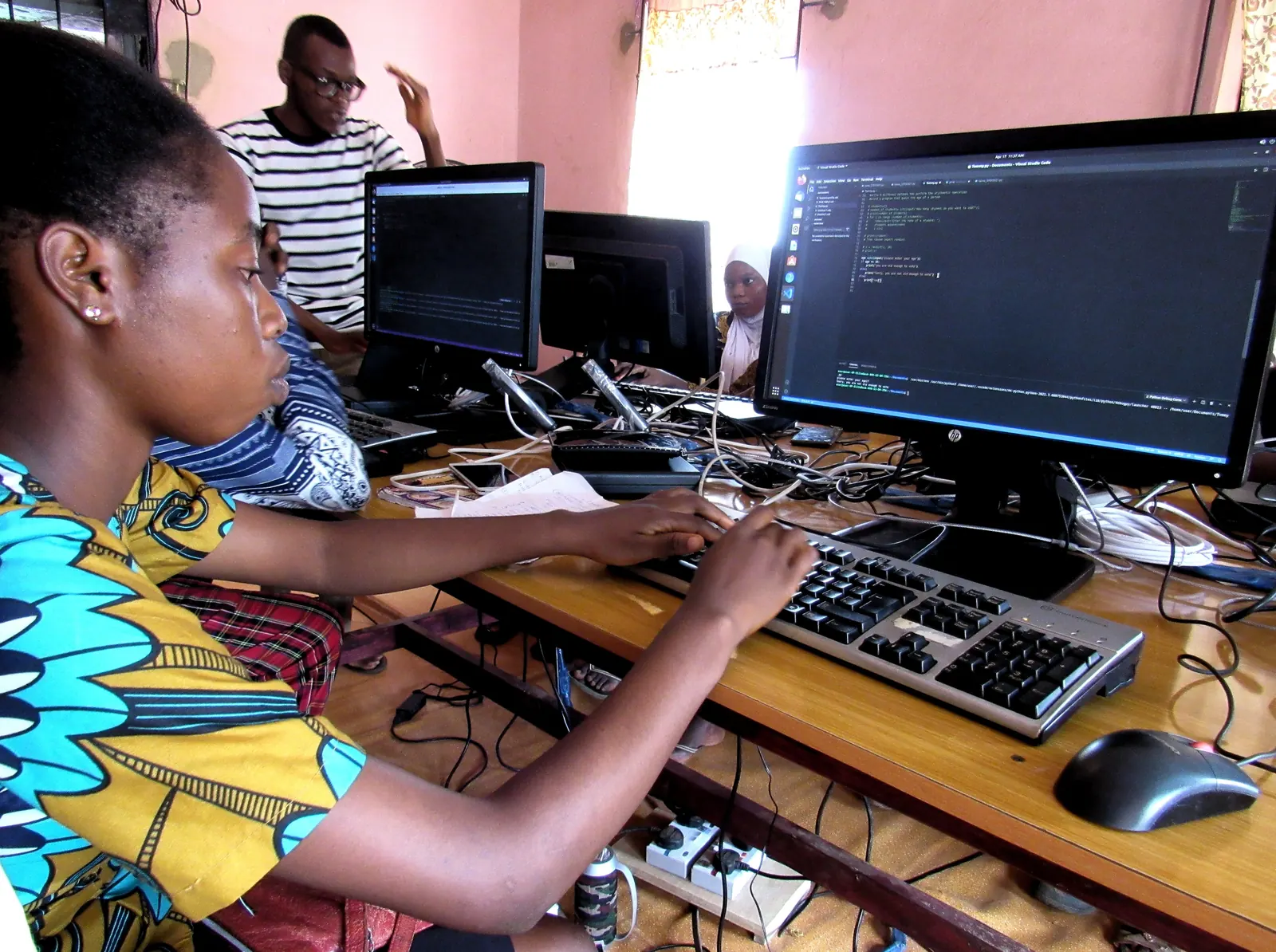 A group of students, most of them young women, gathered around shared computer monitors in a classroom.